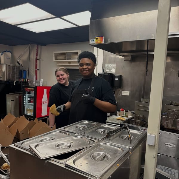 Two smiling Empanada Grill employees in a kitchen, one holding an empanada, the other giving a thumbs up.