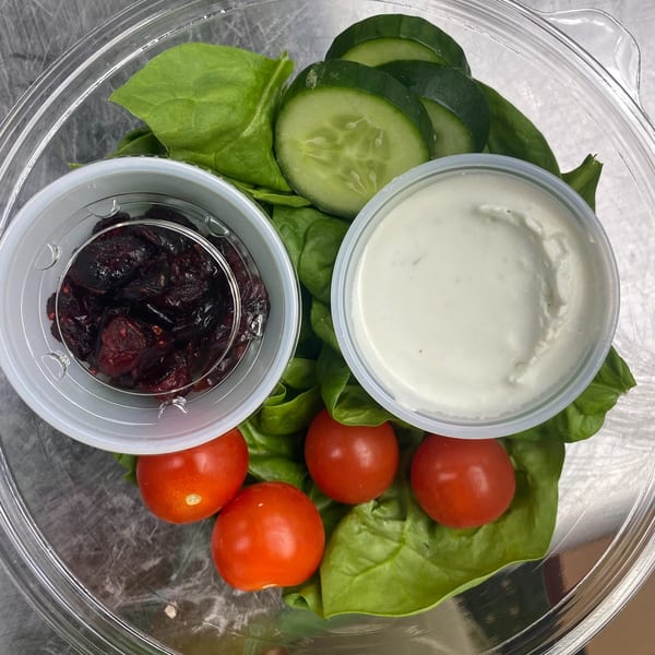 A clear plastic container holds a salad with spinach, cucumbers, cherry tomatoes, dried cranberries, and dressing.