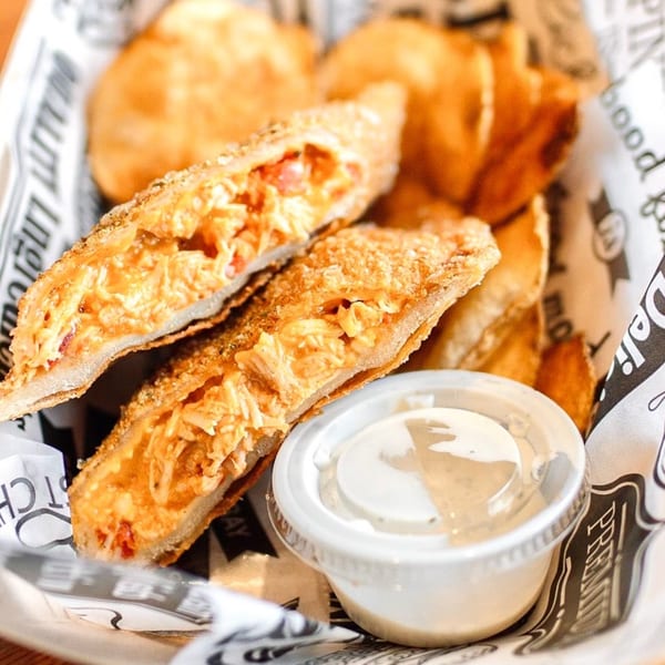 Two golden-brown Empanada Grill empanadas, cut open to reveal a cheesy chicken filling, with a side of dipping sauce.