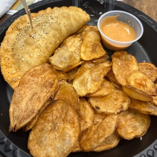 A golden-brown Empanada Grill empanada with seeds, served with crispy potato chips and a side of dipping sauce.